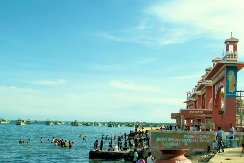 A wide view of pilgrims bathing in the calm blue sea waters at Agni Theertham in Rameshwaram. On the right, a distinctive orange and white temple structure with arched entrances stands along the shoreline, under a bright, clear sky.