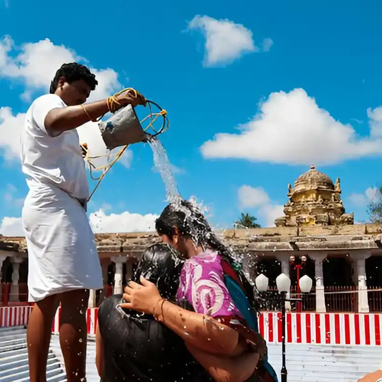 A close-up shot of a temple priest pouring holy water from a silver bucket over a devotee's head at a sacred bath ritual. An ancient stone temple structure stands under a bright blue sky with fluffy white clouds in the background.