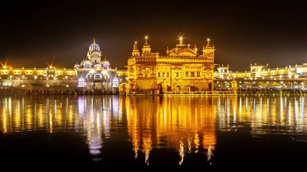 A wide night shot of the illuminated Golden Temple complex in Amritsar. The golden structure and adjacent white marbled buildings cast a brilliant, shimmering reflection across the calm, dark waters of the surrounding sarovar lake.