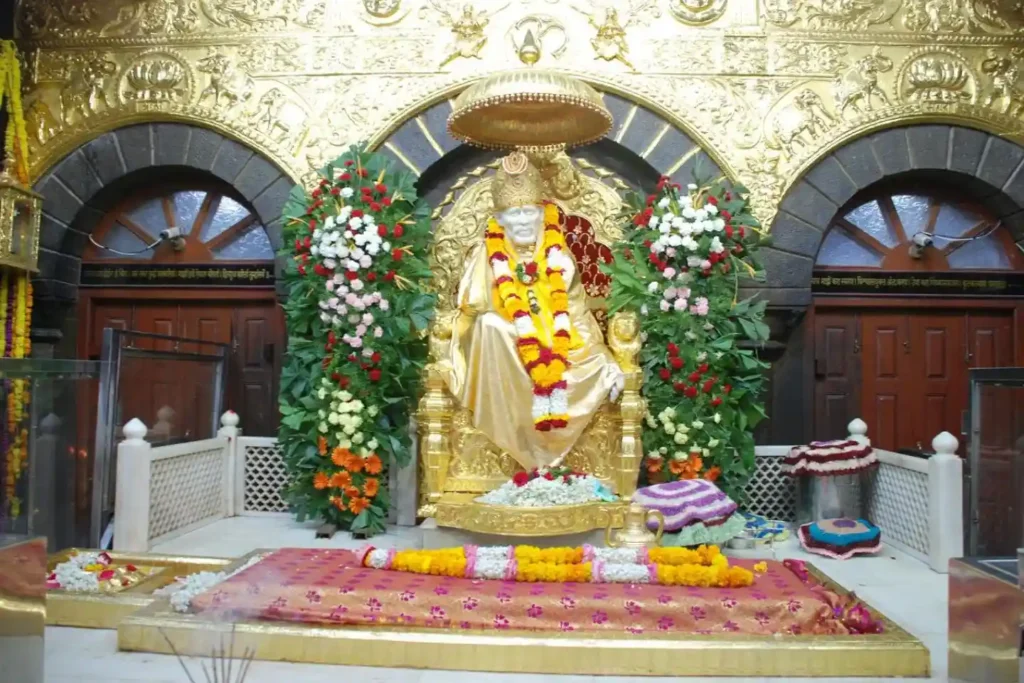 : A vibrant view of the Sai Baba statue in Shirdi, dressed in a bright red shawl and seated on a golden throne, with a long red and yellow floral garland laid over the samadhi structure in the foreground.