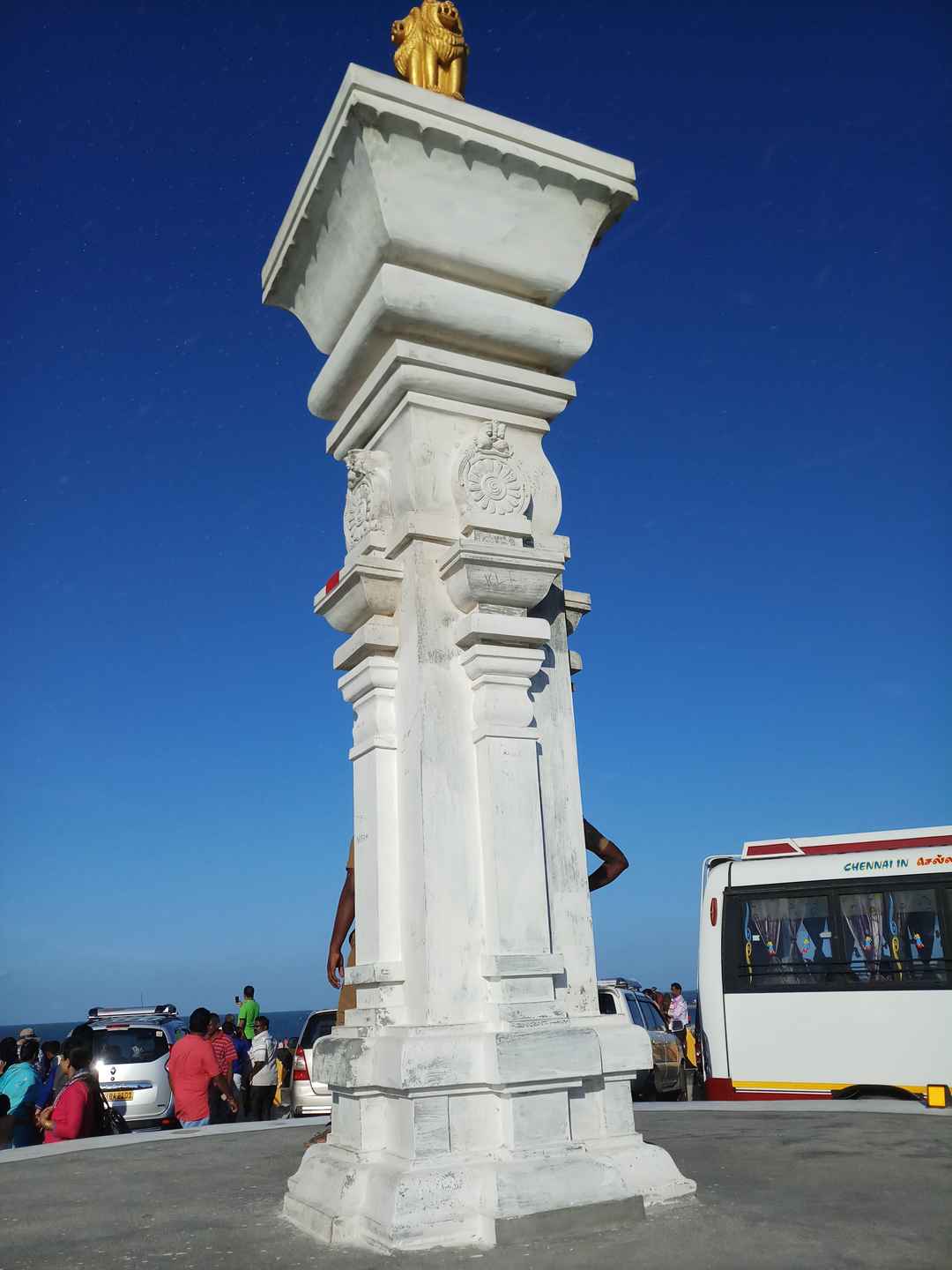 A low-angle shot of a white stone ceremonial pillar standing tall under a clear blue sky. The top of the pillar features a small golden sculpture of the Lion Capital of Ashoka, with people and the side of a white bus visible at its base.
