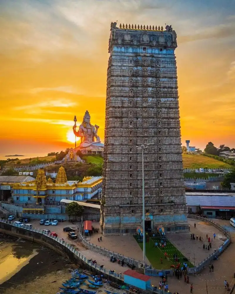 "Towering Murudeshwar temple gopuram beside the giant Shiva statue, silhouetted against an orange coastal sunset"