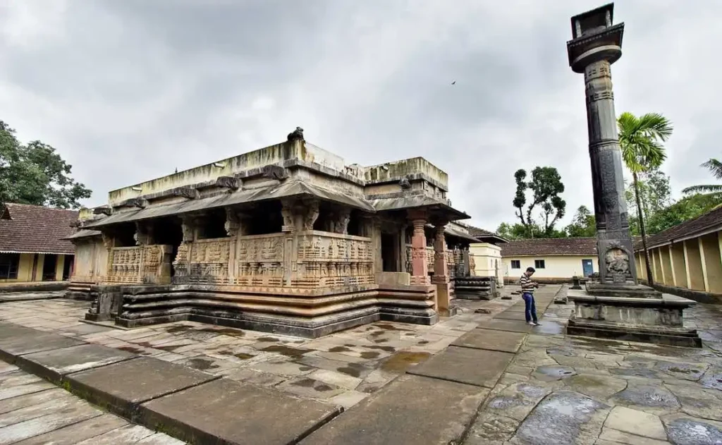 Keladi Rameshwara Temple in Shivamogga, Karnataka—an ancient stone temple courtyard featuring a traditional sloping roof, a tall carved Mahastambha (stone pillar) erected by Rani Chennamma, and stone-paved walkways under a cloudy sky.