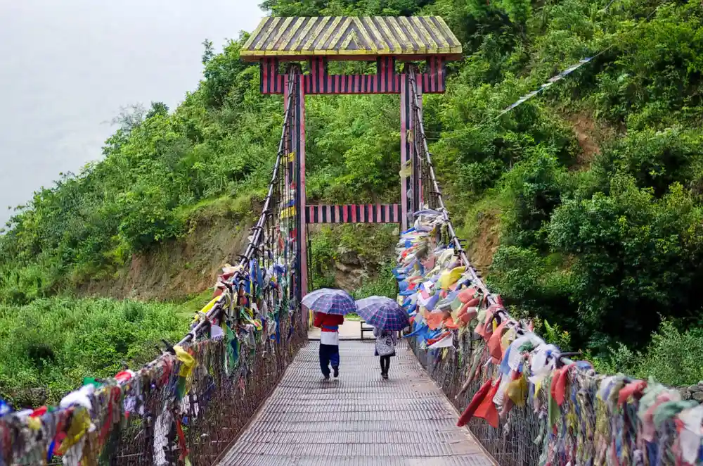 ourists walking with umbrellas on the historic iron suspension Chagzam Bridge draped in Buddhist prayer flags near Tawang, Arunachal Pradesh.